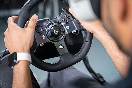 View over the shoulder of a student in front of a steering wheel. Here, the steering of autonomous vehicles is tested (for safety) on the model.