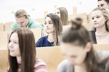 8 Studierende hören konzentriert im Hörsaal zu, während sie an ihrem Platz sitzen und sich teilweise Notizen machen.