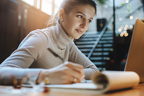 Eine junge Frau sitzt am Laptop und macht Notizen in einem Notizblock.