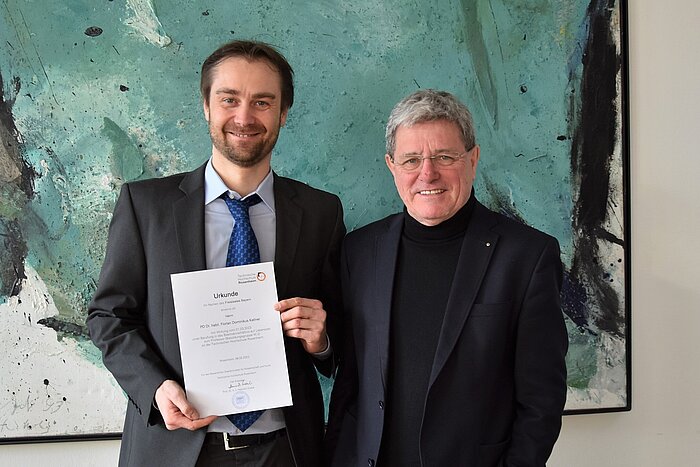 Prof. Dr. Florian Kellner hält seine Vereidigungsurkunde in die Kamera. Er steht neben dem Präsidenten der Hochschule Prof. Heinrich Köster