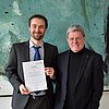 Prof. Dr. Florian Kellner hält seine Vereidigungsurkunde in die Kamera. Er steht neben dem Präsidenten der Hochschule Prof. Heinrich Köster