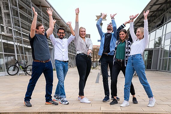 Auf dem Bild sind sechs Studierende der TH Rosenheim zu sehen. Sie befinden sich im Freien, auf der Treppen zwischen R- und S-Bau am Campus Rosenheim. Alle Studierenden heben freudig die Hände in die Höhe und jubeln.