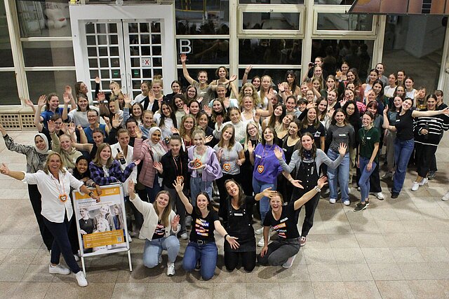 circa 100 Studentinnen stehen passend für ein Foto von oben ausgrichtet und schauen euphorisch, mit in die Luft gestreckte Arme in die Kamera, lachen und tragen normale, ihrem Stil angepasste Kleidung