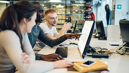  Studierende, die gemeinsam an einem Computer in der Bibliothek recherchieren.