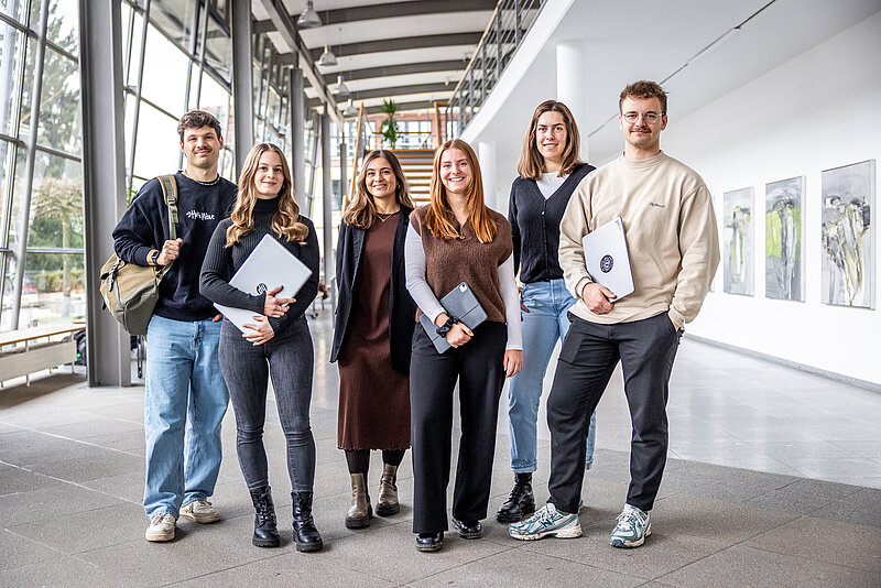 Sechs Studierende stehen nebeneinander im Foyer eines Hochschulgebäudes. Teilweise haben sie Laptops und Tablets in der Hand