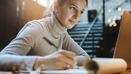 Eine junge Frau sitzt am Laptop und macht Notizen in einem Notizblock.