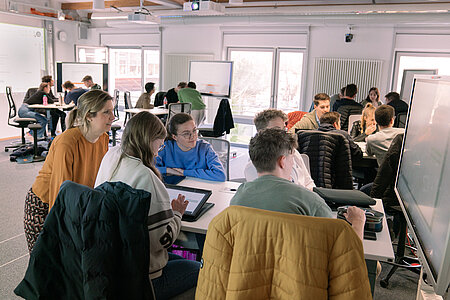 Studierende sitzen im hybriden Lehrraum an einem Tisch mit großem Bildschirm bei der GRuppenarbeit