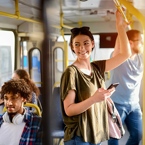 Junge Frau steht im Bus und hält sich am Haltegriff fest. In der anderne Hand hält sie ein Smartphone.