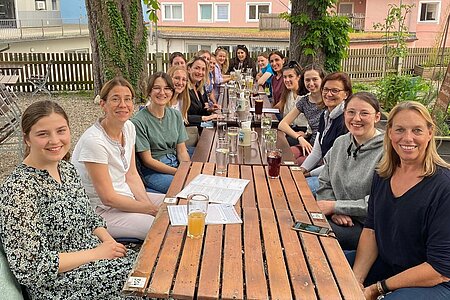 circa 10 Studentinnen und 10 Mentorinnen sitzen gemischt draußen in einem Biergarten an einem Holztisch unter Bäumen. Sie sind sommerlich bekleidet, lächeln freundlich in die Kamera und haben ein Getränk vor sich stehen.