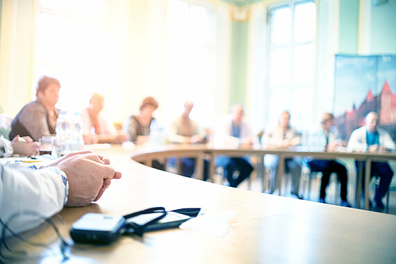 Auf diesem Bild sieht man unscharf mehrere Menschen in einem Besprechungsraum um einen Konferenztisch sitzen.