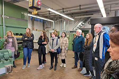 [Translate to English:] Gruppe steht im Holzlabor und hört dem Redner zu.