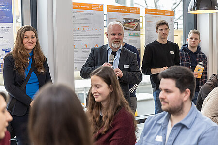 Personen sitzen an Tischen oder stehen bei den Projektplakaten an der Fensterfront des Raumes. Sie hören Prof. Dr. Gerd Beneken während seiner Ansprache an die Teilnehmerinnen und Teilnehmer zu. Der Professor steht dabei im Hintergrund des Bildes, davor sieht man die sitzenden Teilnehmer. 