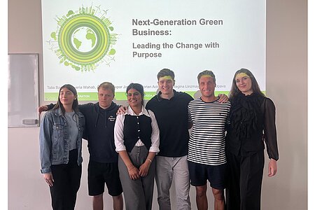 Gruppe aus sechs Studierenden stehen glücklich vor der Leinwand, auf der die Abschlusspräsentation zu sehen ist. Javeria ist unter Ihnen.