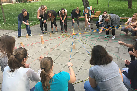 circa 15 Studentinnen hocken draußen auf einem gepflasterten Boden im Kreis. Sie spielen ein Spiel, wo jede das Ende eines Faden in der Hand hat und alle Fäden sich in der Mitte kreuzen. Sie sind sommerlich bekleidet und befinden sich unter Bäumen neben einer grünen Wiese.