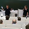 Das Bild zeigt zwei Frauen vor Publikum in einem Hörsaal.