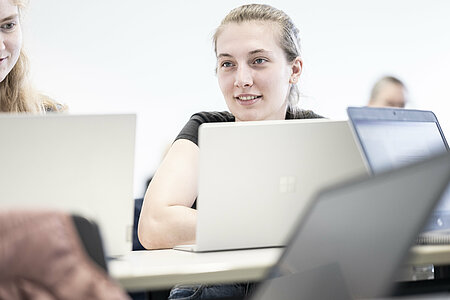 Man sieht 2 Studierende, die humorvoll miteinander kommunizieren, während sie an ihrem Platz sitzen und an Laptops arbeiten.