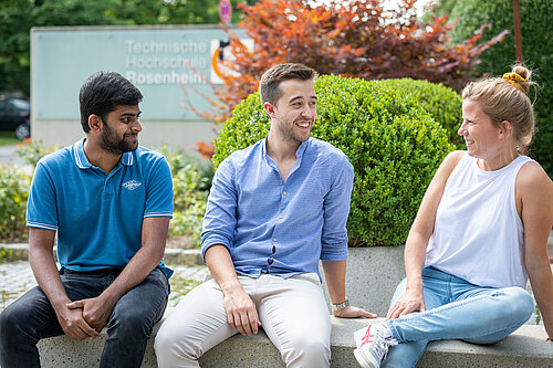 Drei Studen*innen verschiedener Hautfarbe sitzend am Eingangsbereich der Hochschule (Gebäude A).
