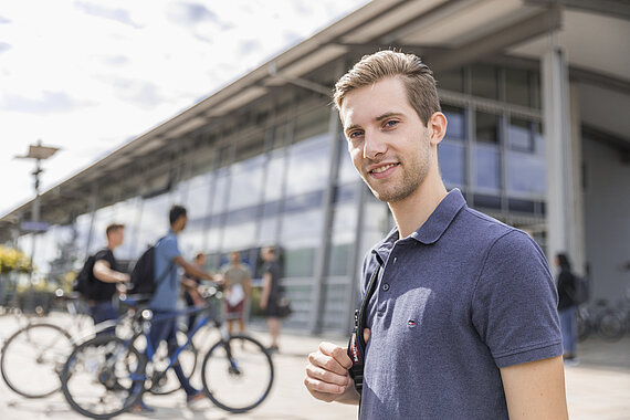 Male student in front of building on Rosenheim with a shoulder bag campus with  