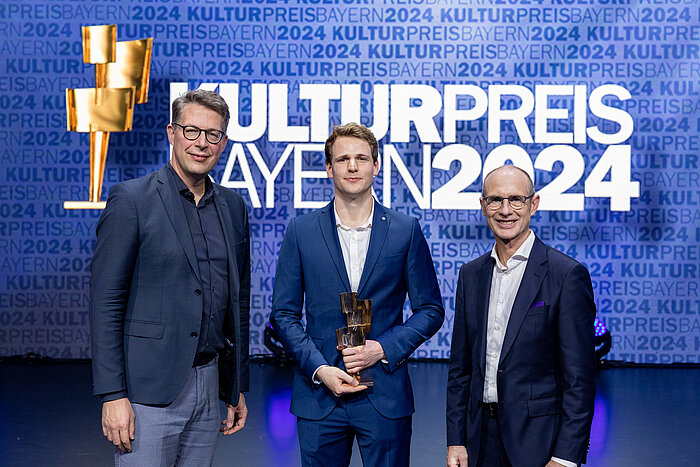 Auf dem Bild sind drei Männer zu sehen. In der Mitte steht der TH-Absolvent Georg Glück. Er wurde mit dem Bayerischen Kulturpreis 2024 ausgezeichnet. Links im Bild ist der Bayerischen Kunst- und Wissenschaftsminister Markus Blume zu sehen. Rechts steht Bayernwerk-Chef Dr. Egon Leo Westphal. 