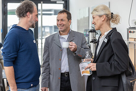 Drei Personen (zwei Männner und eine Frau) stehen lächelnd und mit Getränken in der Hand und sind ins Gespräch vertieft. 