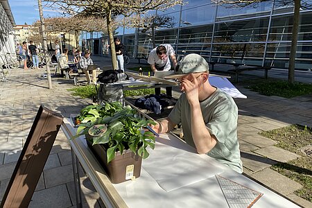 Das Bild zeigt einen Studenten, der im Freien an einem Tisch sitzt und an einem Konstruktionsplan arbeitet.