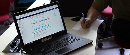 Laptop in working mode, a student sits next to it (you can see a hand with a pen; notes are being taken).