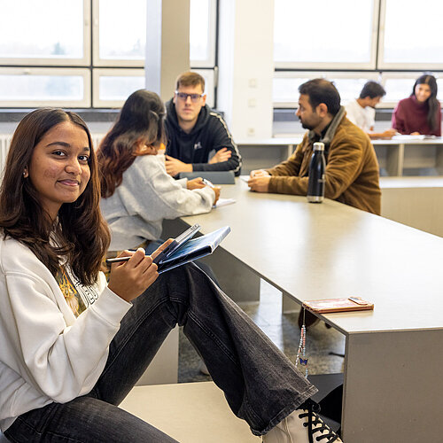 Studentin im Vordergrund sitzt an einer Säule lehnend. Im Hintergrund ist eine Studierendengruppe beim Austausch zu sehen.