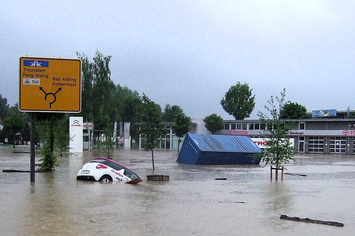 Hochwasser in Rosenheim: überflutete Straße mit Auto und Container vor einem Autohaus