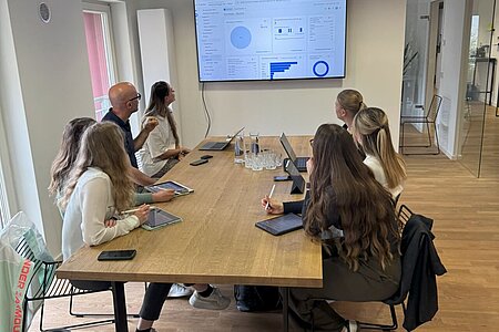 Die Studentinnen sitzen beim Briefing in Büroräumlichkeiten von Bensegger, auf dem digitalen Whiteboard sind Daten und Statistiken zu sehen.