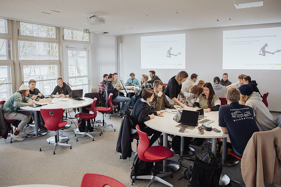 Das Bild zeigt einen großen Raum mit runden Tischen. Studierende arbeiten in Kleingruppen an Aufgabenstellungen.