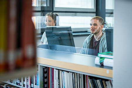 [Translate to English:] Zwei Personen hinter Glas an den PC-Arbeitsplätzen der Bibliothek