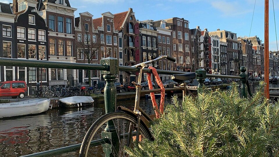 Fürs Praktikum nach Amsterdam Ein Fahrrad lehnt an einem Geländer. Im Hintergrund ist die Kulisse einer Stadt in den Niederlanden.