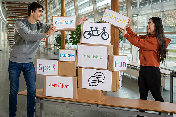 [Translate to English:] Student und Studentin bauen Stapel aus Kartons mit Sprechblasen rund um das Thema Sprache