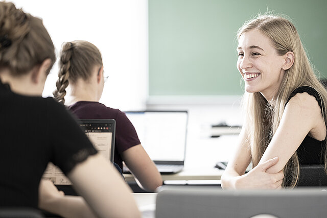 3 Studierende kommunizieren humorvoll miteinander, während sie an ihrem Platz sitzen und an Laptops arbeiten.