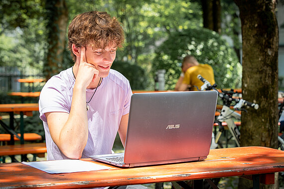 Student sitzt auf Terrasse an Biertisch und arbeitet am Laptop
