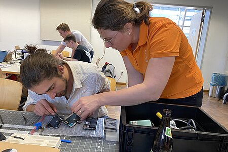 Das Bild zeigt einen Studenten und eine Studentin, die an einem Tisch an einer Leiterplatte arbeiten.