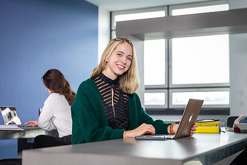 Eine Studentin der TH Rosenheim am Laptop lacht in die Kamara. 