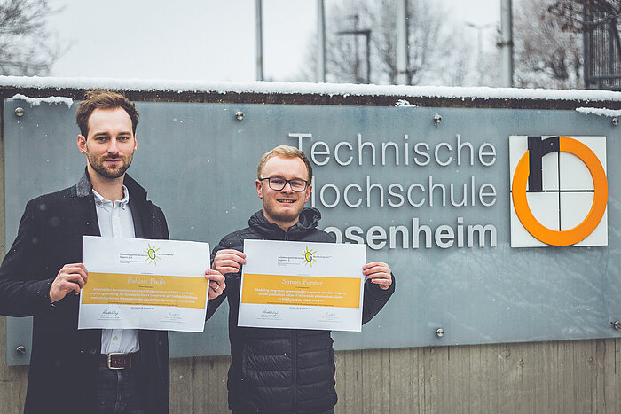 Auf dem Bild sind die beiden ausgezeichneten Hochschulabsolventen Simon Forster (rechts) und Fabian Pauls (links)mit ihren Urkunden zu sehen. Stehen vor dem Logo der TH Rosenheim am Haupteingang.