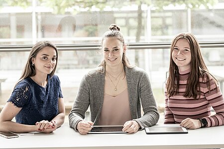 3 Studentinnen sitzen mit 2 Tablets am Tisch. Direkt hinter ihnen ist ein großes Fenster, wodurch man draußen ein anderes Gebäude des Campus sieht.