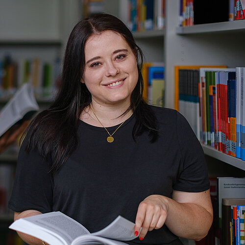 Studentin steht in der Bibliothek neben einem Bücherregal und blättert durch ein Buch