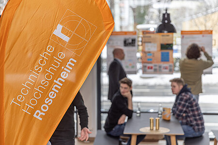 Orangene Beachflag mit dem Logo der TH Rosenheim im Vordergrund. Im Hintergrund sitzen zwei junge Männer an einem Tisch mit Getränken. Dahinter stehen Personen, die sich die Projektplakate an den Fensterscheiben des Raumes ansehen. 