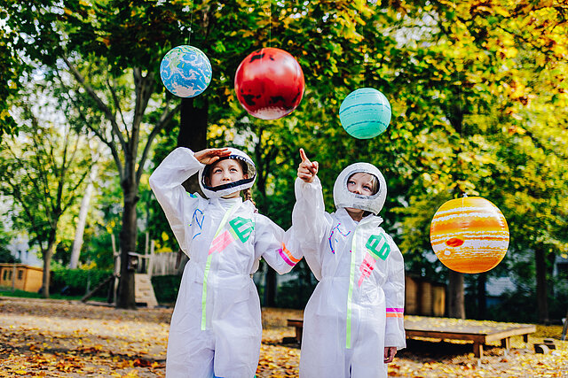 Zwei Kinder haben sich mit Raumanzügen verkleidet, um sie herum schweben gebastelte Planeten