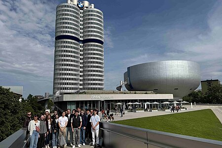 Studierende posieren bei schönstem Wetter vor dem BMW Museum in München. Im Hintergrund ist der BWM-Tower zu sehen.