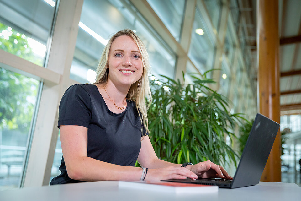 eine studentin mit dunklem Oberteil und blonden Haaren arbeitet am Laptop und schaut dabei in die Kamera