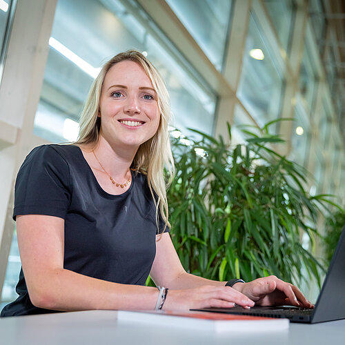eine studentin mit dunklem Oberteil und blonden Haaren arbeitet am Laptop und schaut dabei in die Kamera