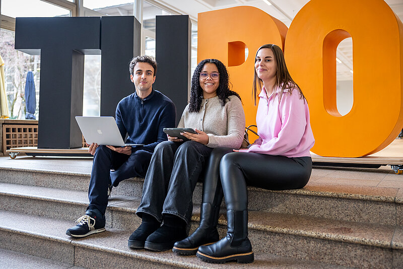 Zwei Studentinnen und ein Student sitzen auf Treppenstufen. Hinter ihnen stehen große Buchstaben T, H, R und O.