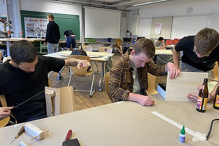 Das Bild zeigt drei Studenten, die in einem großen Raum sitzen und an verschiedenen Gegenständen aus Holz arbeiten.