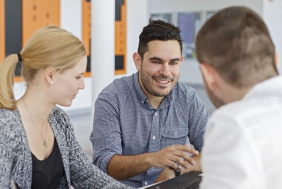 Eine Studentin und zwei Studenten gegenübersitzend an einem Tisch