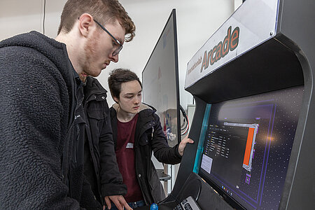 Zwei junge Männer (ein weiterer verdeckt zu sehen) stehen an einer eigens gebauten Computerspiel-Station mit Joystick und spielen. 