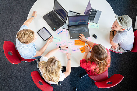 Vier Studenten hocken mit ihren Laptops und Schreibutensilien um einen runden Tisch.